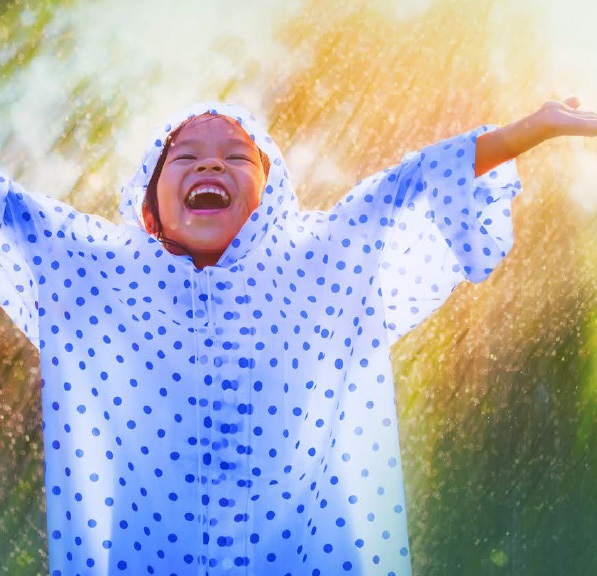 Smiling child in a raincoat standing with arms outstretched in the sun and rain