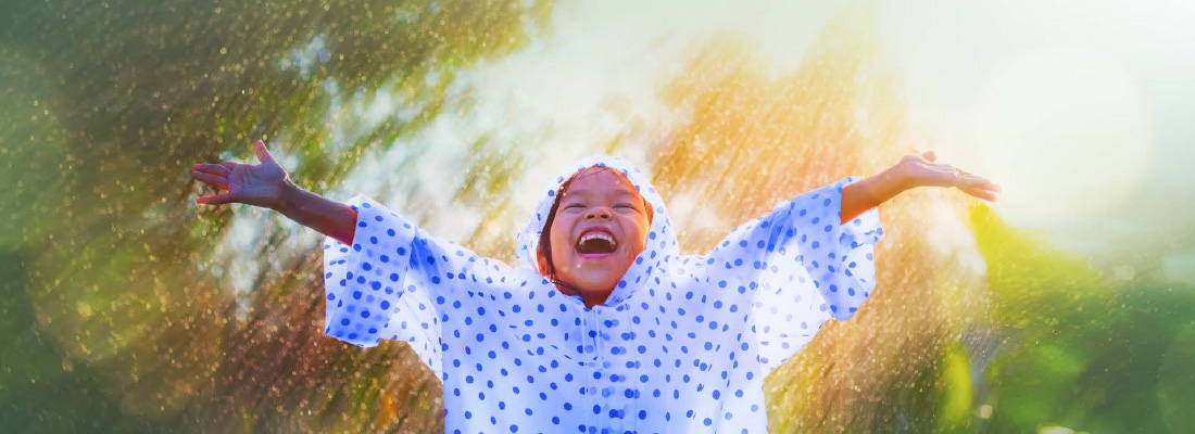 Smiling child in a raincoat standing with arms outstretched in the sun and rain