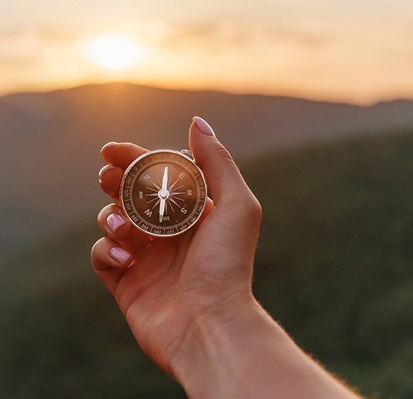 A hand holds a compass towards the sun rising over a mountain