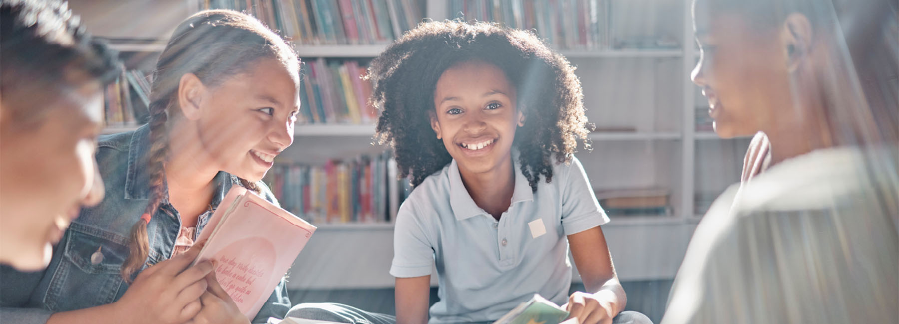 Four young people sit in a circle in a library holding books and smiling