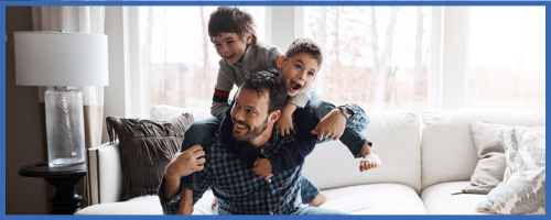 A smiling man sits on a sofa with two joyful young boys on his back