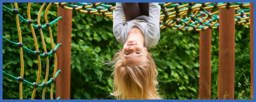 A young child hangs upside down by their knees from a rope climbing structure outdoors
