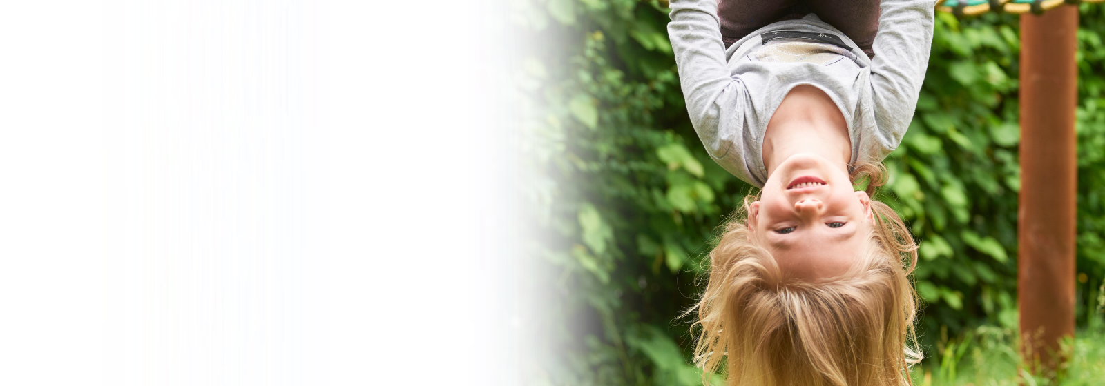 A young child is smiling while hanging upside down from a rope net climber outdoors