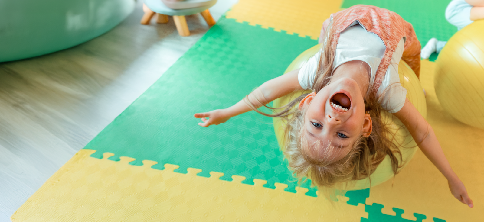 A child with a big happy smile is upside down on a yellow exercise ball
